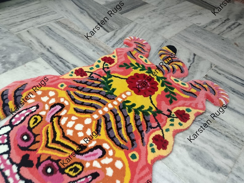 May include: A colorful rug shaped like a tiger with pink, yellow, orange, black, and green accents. The rug has a floral design with a large pink flower in the center. The rug is made of wool and is hand-knotted. The rug is made by Karsten Rugs.