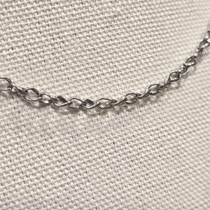 May include: A silver-toned metal chain necklace featuring a series of interconnected oval-shaped links. The chain has a polished, reflective finish and is displayed in a curved, horizontal orientation against a textured, off-white fabric surface.