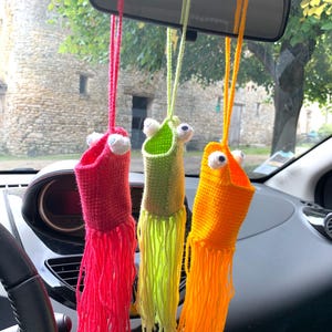 Könnte beinhalten: Drei farbenfrohe, handgefertigte Häkel-Autoanhänger, die an einem Rückspiegel hängen. Die Anhänger sind rot, grün und orange, jeweils mit Fransen unten und Wackelaugen. Der Innenraum des Autos ist im Hintergrund zu sehen.