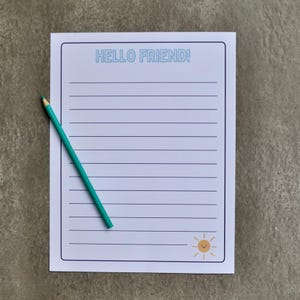 May include: A lined notepad with the text "HELLO FRIEND!" in light blue. A small, smiling sun graphic is in the bottom right corner. A teal pencil rests diagonally across the notepad.