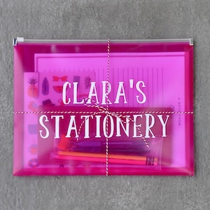 May include: A vibrant pink stationery set in a clear plastic zip-up pouch, tied with white and red twine. The text "Clara's Stationery" is printed in white. The set includes paper, pencils, and stickers.