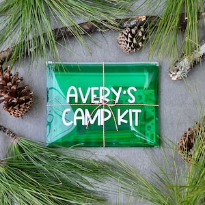 May include: A green cellophane package tied with twine, labeled "AVERY'S CAMP KIT" in white lettering. The package is surrounded by pine needles and pine cones, suggesting a camping or outdoor theme.