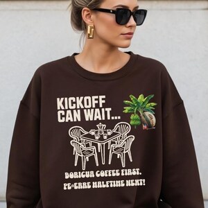 May include: A brown sweatshirt with white text that reads "Kickoff Can Wait..." above a graphic of a table with chairs and coffee cups. Below the graphic, the text reads "Boriqua Coffee First. Pe-erre Halftime Next!" A small graphic of a palm tree and football is on the right.