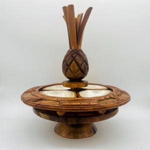 May include: A wooden serving dish with a carved pineapple topper. The dish has a round base and a tiered design with four white compartments. The outer rim is decorated with carved details, showcasing a natural wood finish.