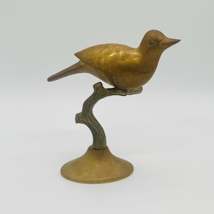 Puede incluir: Figura de pájaro de latón dorado, posada en una rama, montada sobre una base redonda. El pájaro es de color oro cálido, con una forma detallada. La rama y la base tienen un aspecto más oscuro y envejecido. Un acento decorativo para la decoración del hogar.