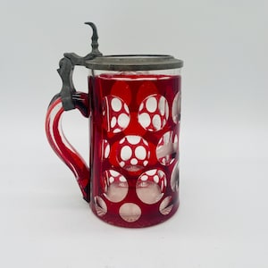 Puede incluir: Una jarra de cerveza de cristal rojo rubí con tapa y asa metálicas con bisagras. El cristal presenta un patrón recortado de círculos blancos. El asa también es roja y blanca. La jarra es de diseño clásico.