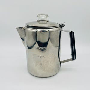 May include: A stainless steel coffee percolator with a clear glass knob on the lid. The pot has a black handle and the text "NINE CUPS" is visible. Measurement markings for 5 and 7 cups are also present.