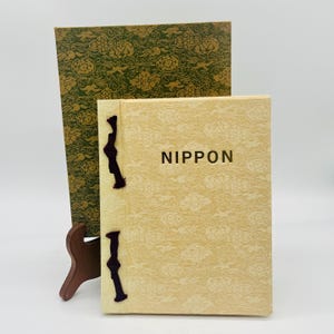 Op de afbeelding: Een vintage boek getiteld "NIPPON" met een crèmekleurige omslag met een wolkenpatroon. Het boek is gebonden met donkerpaarse banden. Achter het boek staat een groene boekomslag met een gouden bloemenmotief.