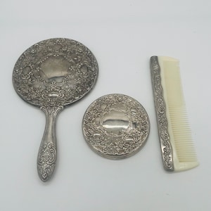 May include: A vintage vanity set comprising a hand mirror, a compact, and a comb. The silver-toned items display intricate floral patterns. The mirror is oval with a handle. The compact is round, and the comb has a cream-coloured base.