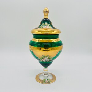 May include: A decorative green and gold glass jar with a lid. The jar features gold bands and floral accents. The lid is topped with a gold finial. The jar sits on a clear glass stem and a gold base.
