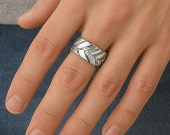 Könnte beinhalten: Ein silberner Ring mit einem Zickzackmuster. Der Ring wird an einem Finger getragen und zeigt sein geometrisches Design. Der metallische Glanz des Rings kontrastiert mit dem Hautton und dem Jeans-Hintergrund.