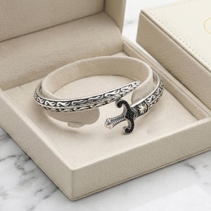 Puede incluir: Una pulsera plateada con un diseño ornamentado, con un cierre en forma de espada con detalles negros, descansa en una caja de joyería beige abierta. Se ve una caja a juego con un logotipo dorado. Se destacan los intrincados detalles de la pulsera.