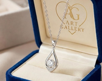 May include: A silver pendant necklace with a teardrop design, featuring a large, round, clear gemstone at the center, surrounded by smaller stones. The necklace is displayed in a blue velvet jewelry box. The box has the text "AG ART GALLERY".