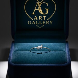 Puede incluir: Un anillo de plata con la letra "A" en cursiva, presentado en un joyero de terciopelo azul abierto. La tapa del joyero muestra el texto dorado "AG ART GALLERY" dentro de un corazón. Una joya delicada.