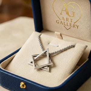 May include: A silver-toned necklace with a Star of David pendant, presented in a blue jewelry box with a cream-colored interior. The box is embossed with "AG ART GALLERY" in gold lettering. The pendant is geometric and modern.