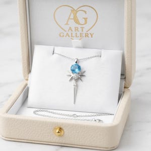 May include: A silver necklace with a light blue gemstone pendant in an open beige jewelry box. The pendant has a starburst design with a long, pointed element. The box has the text "ART GALLERY".