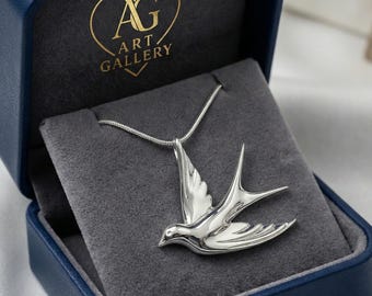 May include: A silver-colored bird pendant necklace displayed in a blue jewelry box with a gray velvet interior. The pendant is a detailed representation of a bird in flight. The box has a gold logo that says "AG ART GALLERY".