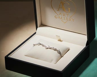 May include: A silver bracelet with a leaf design, set with small clear stones, is displayed on a white cushion within a black jewellery box. The box is lined with cream fabric and has gold text: "AG ART GALLERY".