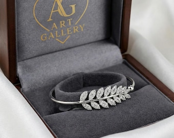 May include: A silver bracelet with a leaf design, set with clear gemstones, is displayed in a wooden jewellery box. The box has a grey velvet interior and gold lettering that reads "AG ART GALLERY".