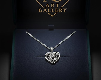 May include: A silver heart-shaped pendant necklace with an intricate openwork design, displayed in a dark blue jewelry box. The necklace is on a silver chain. The box has a gold logo that says "AG ART GALLERY".