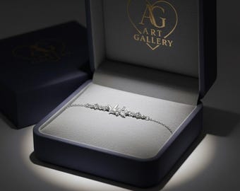 May include: A silver bracelet with a butterfly design, presented in an open navy blue jewellery box. The box has a grey interior and the words "AG ART GALLERY" in gold lettering. The bracelet is delicate and features small, sparkling stones.