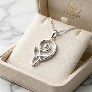 May include: A silver pendant necklace with a Celtic knot design, displayed in a cream-colored jewelry box. The necklace features a detailed, swirling pattern and is presented against a soft, padded interior. The box has a gold logo.