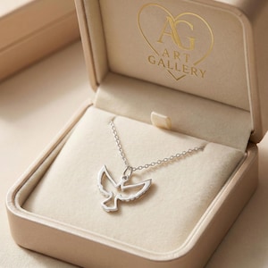 May include: A silver dove pendant necklace presented in a beige jewellery box. The necklace has a fine chain and a dove-shaped pendant. The jewellery box has the text "ART GALLERY" and a heart-shaped logo.