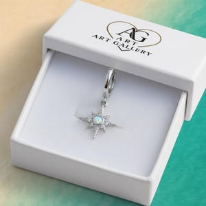 May include: A silver-colored starburst charm with a central opal-like stone and surrounding clear crystals, suspended from a hoop. The charm is presented in a white box with the "AG ART GALLERY" logo.