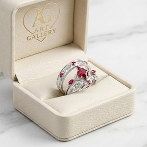 May include: A silver ring set with red gemstones and clear accent stones, displayed in an open cream-colored jewelry box. The box has the text "AG ART GALLERY" in gold lettering. The ring set includes a square-cut center stone.