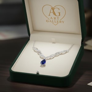 Pode incluir: Um colar de prata com um pingente em forma de gota, com uma pedra preciosa azul escura, exibido em uma caixa de joias verde. O colar tem um design delicado com pequenas pedras transparentes. A caixa está aberta, revelando um interior creme e o texto "AG ART GALLERY".
