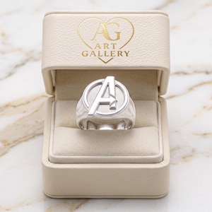 May include: A silver-toned ring featuring a prominent letter A inside a circular frame, presented in a beige rectangular jewelry box. The interior of the box lid displays a gold-colored heart logo with the text AG ART GALLERY printed inside.
