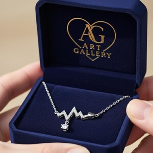Op de afbeelding: Een zilveren ketting met een hartslagontwerp, gepresenteerd in een donkerblauwe fluwelen sieradendoos. De ketting heeft een hanger met een kleine, heldere edelsteen. De doos heeft de tekst "AG ART GALLERY" in goud.