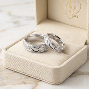Könnte beinhalten: Zwei silberne Ringe mit Blattmuster liegen in einer offenen, cremefarbenen Ringschatulle. Die Schatulle befindet sich in einer größeren cremefarbenen Schachtel mit den Worten "AG ART GALLERY" in goldfarbenen Buchstaben. Der Hintergrund ist eine Marmoroberfläche.