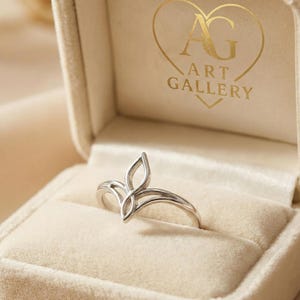 Peut inclure: Une bague argentée avec un motif en forme de feuille, présentée dans un écrin de bijoux en velours beige. La bague présente un design ajouré avec deux formes incurvées et pointues. La boîte porte l'inscription "AG ART GALLERY" en lettres dorées.