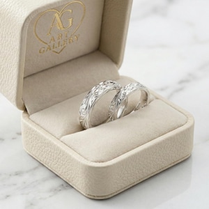 May include: Two silver rings with detailed floral designs are displayed in an open beige ring box. The box's interior is a soft cream colour. The box has the text "ART GALLERY" in gold lettering.