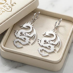 May include: Silver dragon earrings displayed in a cream-colored jewelry box. The earrings feature a detailed dragon design with intricate cutouts. The box is open, revealing the earrings against a cream-colored interior. The box lid has the text "ART GALLERY".