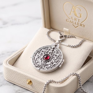Peut inclure: Un pendentif médaillon circulaire argenté orné de volutes en relief complexes et d'une pierre précieuse rouge ronde au centre. Le médaillon est suspendu à une chaîne argentée et présenté dans un écrin crème portant le texte AG ART GALLERY.