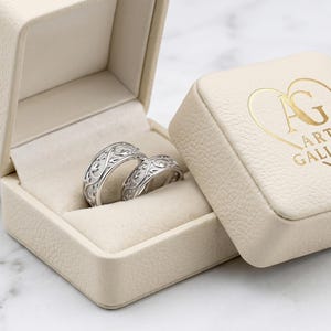 Puede incluir: Dos anillos de plata con diseños intrincados descansan en un joyero crema abierto. La caja está junto a una caja cuadrada a juego con el texto dorado "AG ART GALLERY" y un logotipo de corazón. Los anillos están sobre una superficie de mármol.