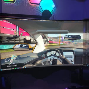 Può includere: Una persona sta giocando a un videogioco di corse su un grande monitor curvo. Il gioco mostra una visuale in prima persona di un'auto che guida su un circuito di gara. L'auto è gialla e ha un volante nero. Il cruscotto mostra un tachimetro che segna 000.