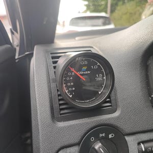 Może przedstawiać: Czarny i srebrny wskaźnik doładowania turbo zamontowany na desce rozdzielczej samochodu. Wskaźnik pokazuje odczyt 0,2 na tarczy. Wskaźnik jest oznaczony "GReddy" i "Turbo Boost". Deska rozdzielcza jest czarna.