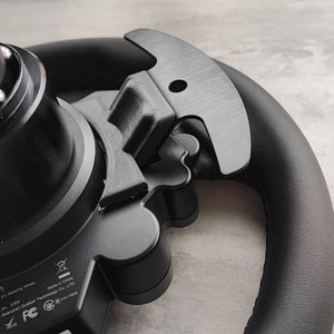 Puede incluir: Primer plano de un volante negro con un accesorio de plástico negro. El accesorio tiene un agujero en el centro y una forma curva. El volante tiene una cubierta de cuero negro y el texto "ES Steering Wheel", "Made in China", "Shenzhen Gudsen Technology Co., LTD", "FC" y "3.6W" es visible en el volante.