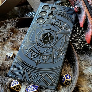 May include: A black phone case with a detailed geometric design featuring a large, silver-colored die in the center. The case is surrounded by dice on a furry surface.