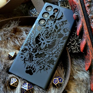 Puede incluir: Una funda de tel&eacute;fono negra con un dise&ntilde;o detallado de un ciervo, un lobo y otros animales en un entorno de bosque. La funda tiene una protuberancia de c&aacute;mara elevada con tres lentes.