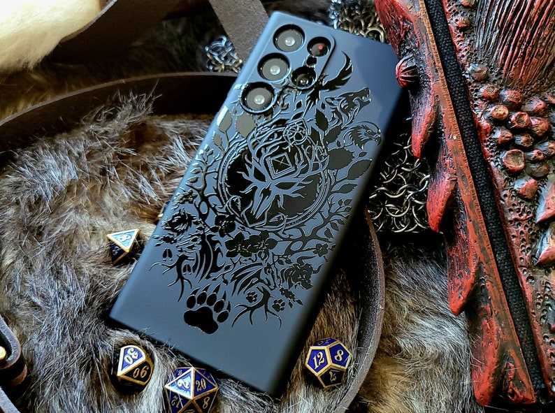 Puede incluir: Una funda de tel&eacute;fono negra con un dise&ntilde;o detallado de un ciervo, un lobo y otros animales en un entorno de bosque. La funda tiene una protuberancia de c&aacute;mara elevada con tres lentes.