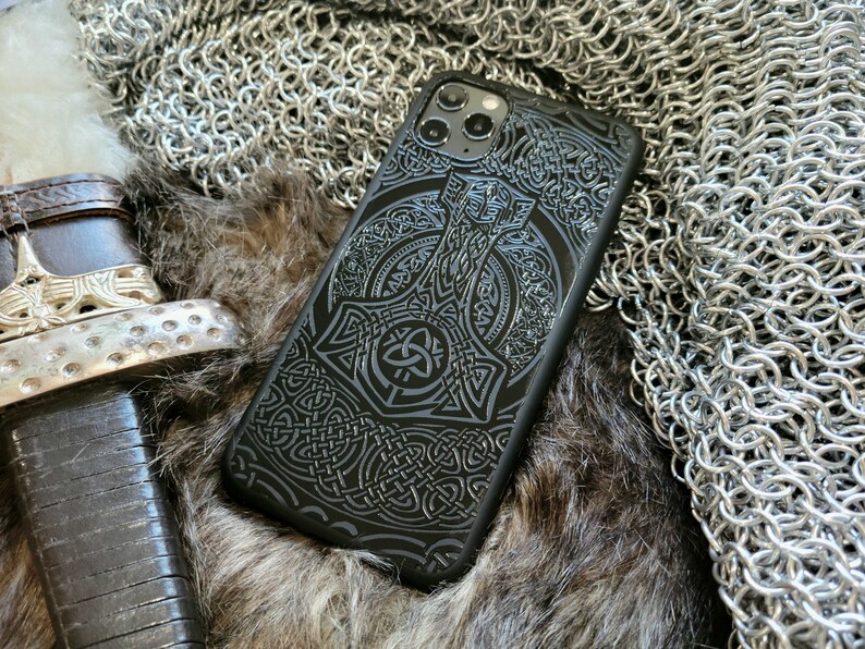 May include: Black phone case with a detailed design of a hammer and intricate knotwork. The case has a raised camera bump with three lenses.