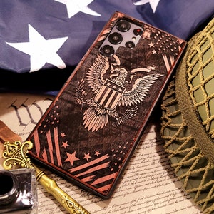 Pode incluir: Uma capa de telefone preta com um padrão de grão de madeira e um design de bandeira americana vermelho, branco e azul. A capa apresenta uma imagem esculpida de uma águia com um escudo e estrelas. A capa do telefone está sobre uma superfície branca com uma pena e um tinteiro.