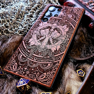 May include: A wooden phone case with an intricate engraved design featuring a central cross, wings, and circular patterns. The case is brown with a lighter engraved design. Several blue and gold dice are scattered around the case.