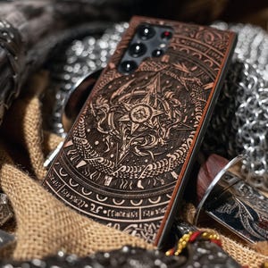 May include: A brown wooden phone case with an engraved design of a compass rose, serpents, and symbols. The case is surrounded by chainmail, a horn, and a sword, suggesting a fantasy theme.