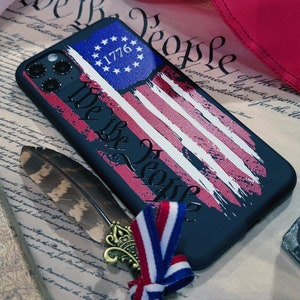 May include: A black phone case with a distressed American flag design. The flag has red and white stripes and a blue canton with 13 white stars and the text "1776". The text "We the People" is written in a distressed font across the flag.