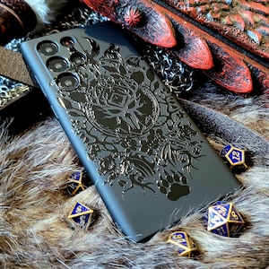 Puede incluir: Una funda de tel&eacute;fono negra con un dise&ntilde;o detallado de un &aacute;rbol y otros elementos de la naturaleza. La funda est&aacute; rodeada de dados azules y dorados sobre una superficie peluda marr&oacute;n.